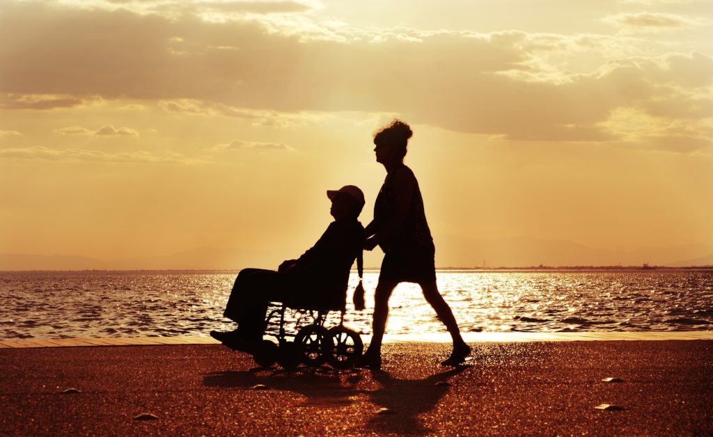 Eldre kvinne som triller eldre mann i rullestol på stranden i solnedgang