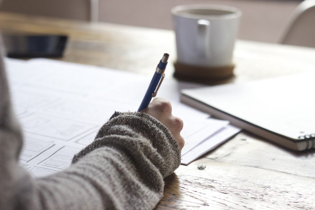 Bilde av en høyre hånd som skriver med penn på dokumenter. Det er en kaffekopp på bordet.